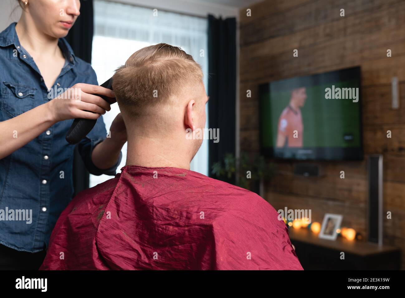 Wife cutting husband's hair at home in front of TV with clipper and holding comb in other hand. Stock Photo