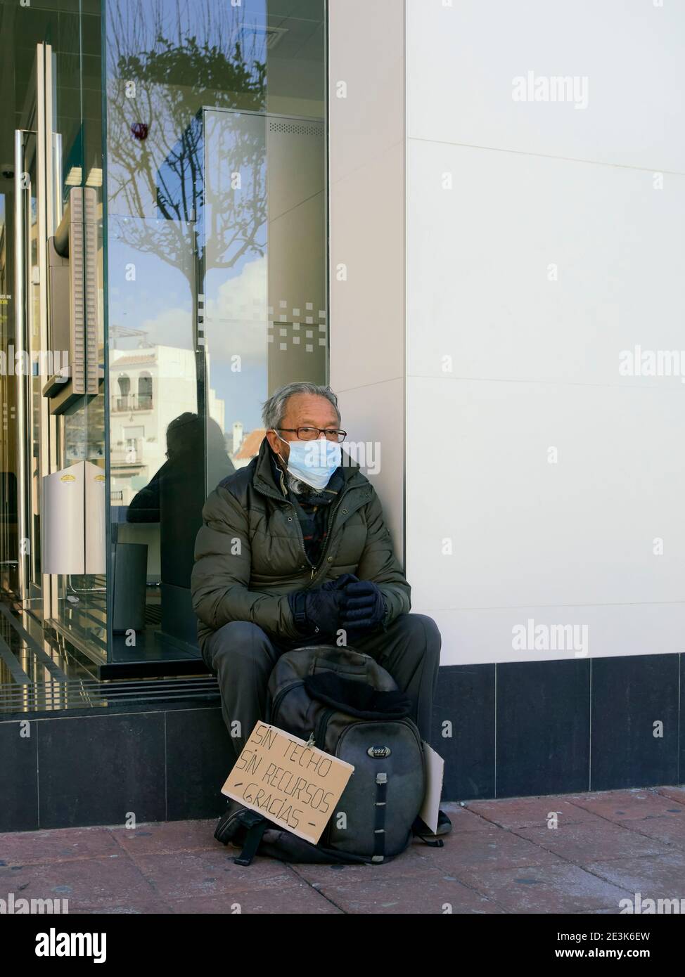 An elderly homeless Spanish man wearing a surgical mask begs outside a bank during the Corornavirus pandemic, Nerja, Malaga, Spain Stock Photo