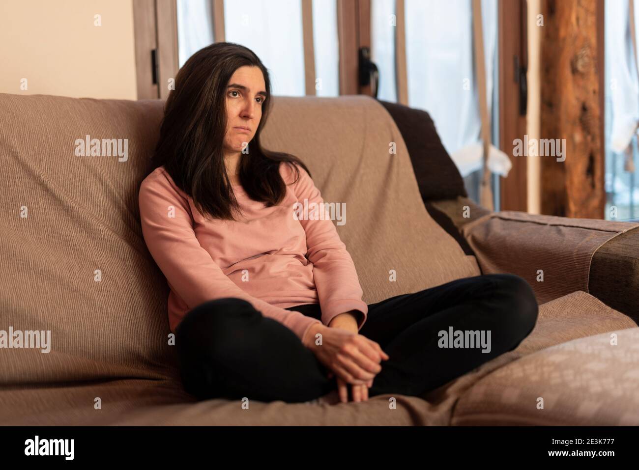 sad happy brunette woman sitting on a sofa in tracksuit with crossed legs looking at TV Stock Photo