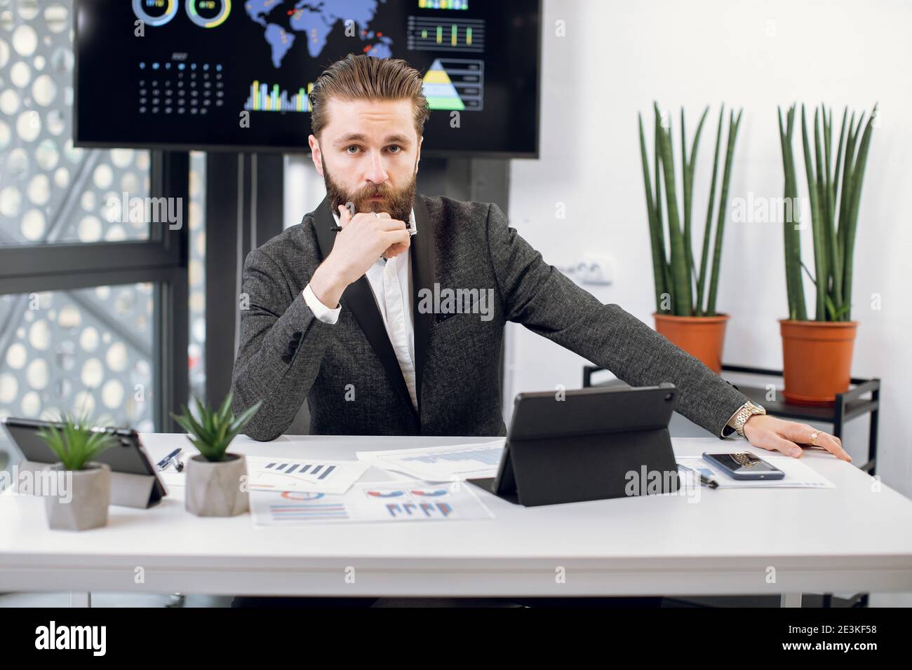 Handsome purposeful confident bearded businessman, chief executive officer in formal wear, sitting at workplace and looking at camera. Male worker Stock Photo
