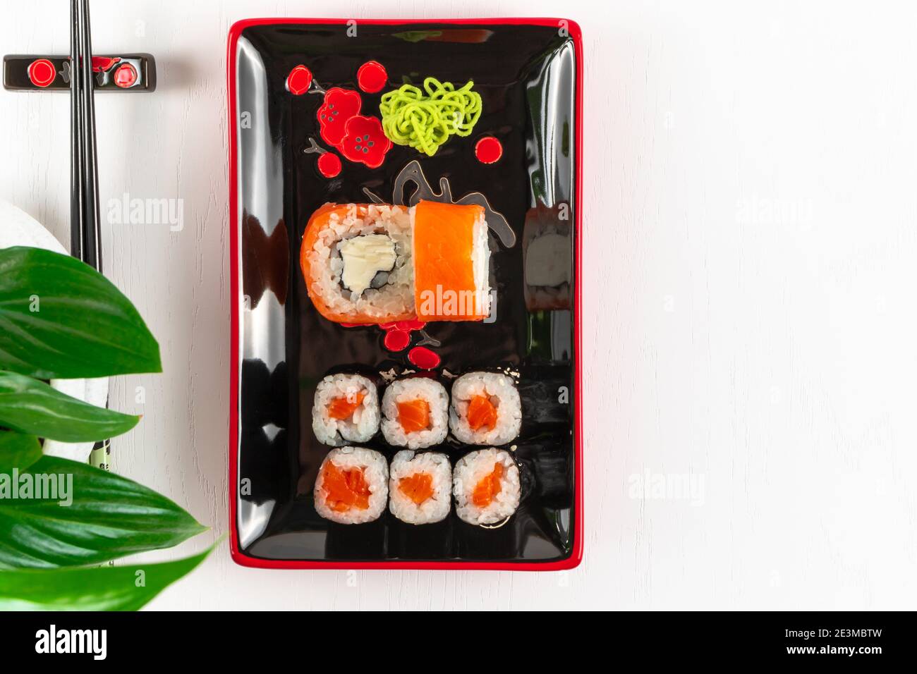 fresh appetizing rolls with salmon in nori seaweed in black-red plates on a white plate. near young green leaves of a plant Stock Photo