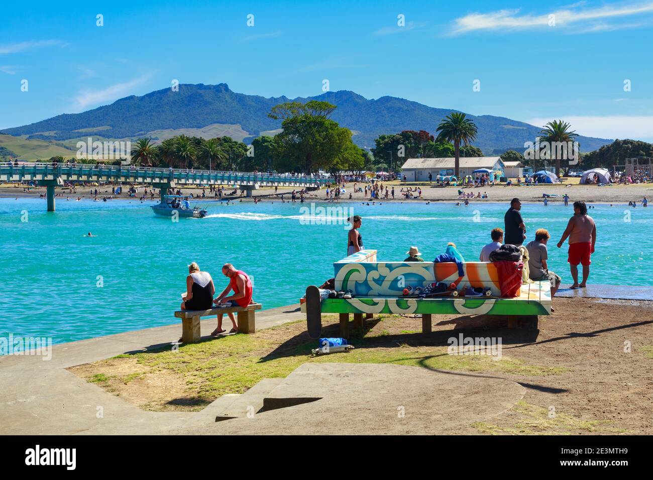 People on the waterfront in Raglan, a seaside town in New Zealand, on a hot summer's day. To the left, Te Kopua footbridge crosses the harbor Stock Photo
