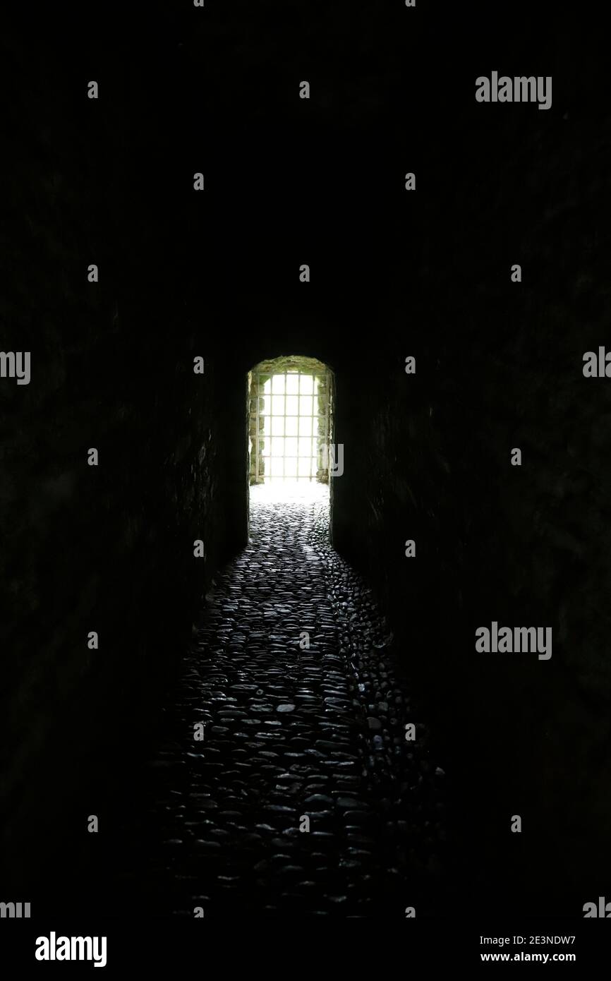 Dramatic, dark, stone paved tunnel ending in bright opening blocked by metal grate Stock Photo