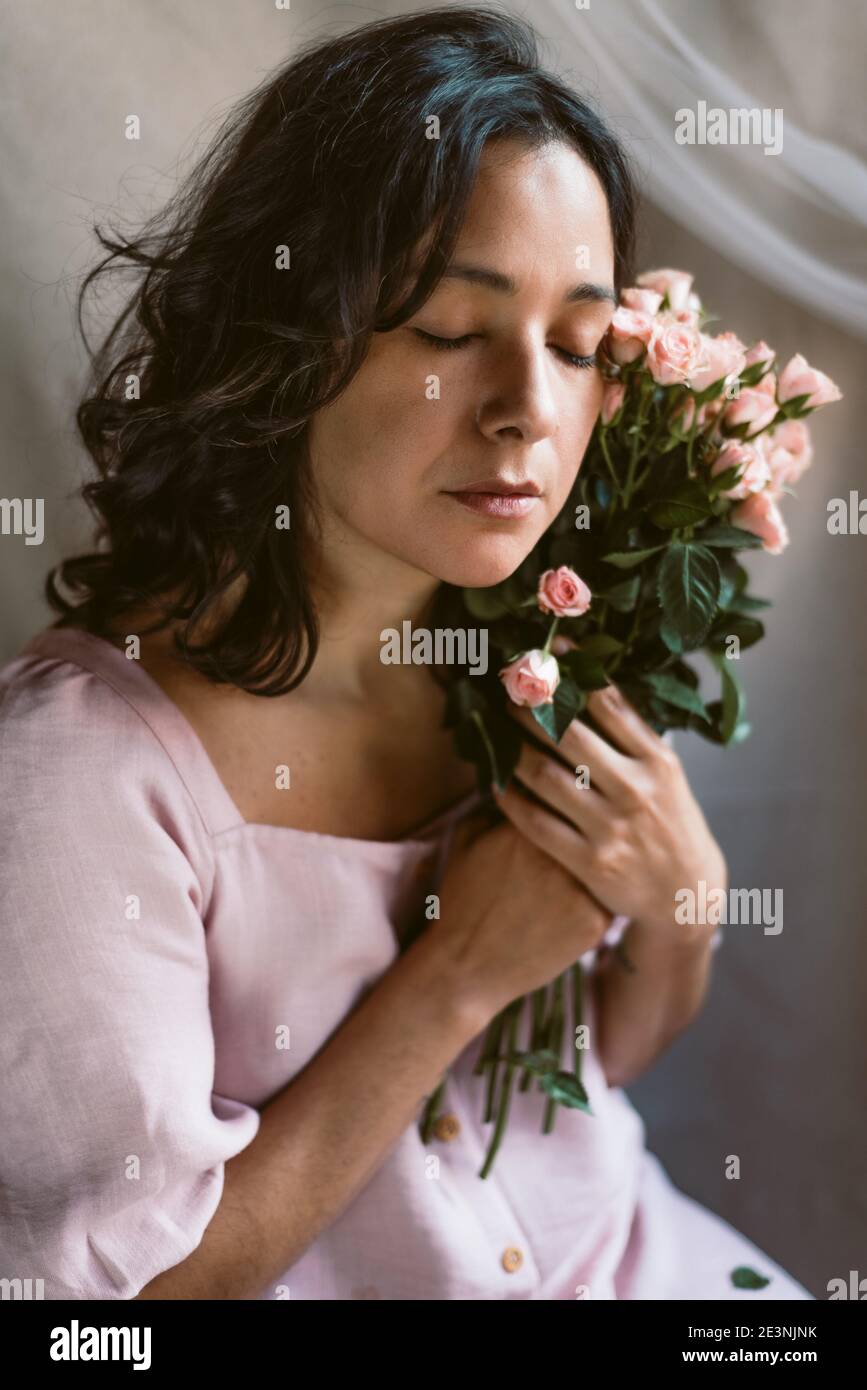 elegant woman portrait with pink roses and pink dress and natural look vertical close up holding flowers bouquet. closed eyes. midlife fragile woman. Stock Photo