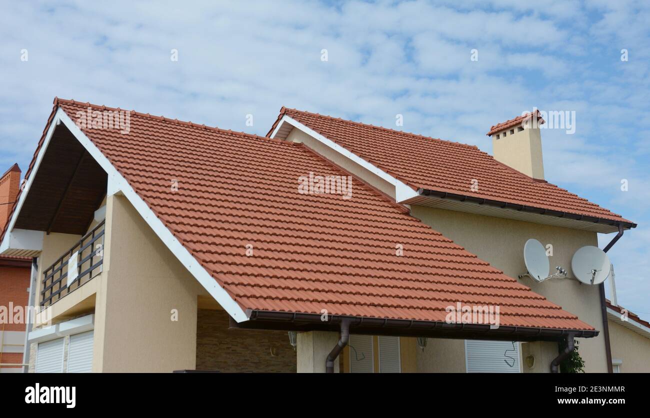 A close-up of stucco house with red metal tiled double gabled roof, a chimney, fascia boards, rain gutters, two satellite dish antennas and an attic. Stock Photo