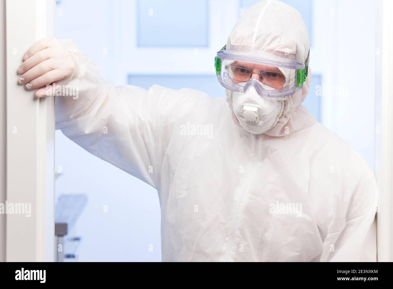 Tired doctor because of covid-19 standing in white protective clothing and medical mask in front of a surgery in a hospital - focus on the face Stock Photo