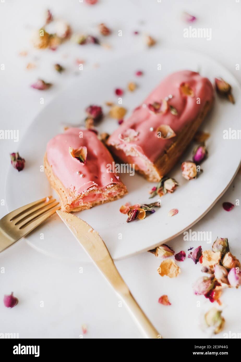 Rose eclair dessert with dried rose petals on white oval plate with golden cutlery over white marble background, selective focus. Artisan homemade desserts concept Stock Photo