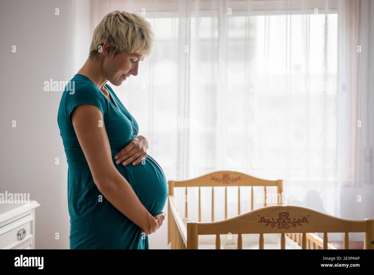 Pregnant woman holding her belly by baby cot next to the window Stock Photo