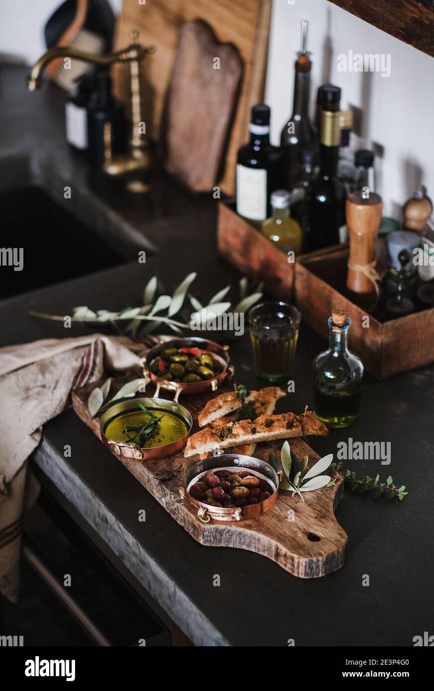 Pickled Greek olives, olive oil in copper jars and herbed focaccia slices on rustic wooden board over kitchen counter. Traditional Mediterranean meze appetizers platter Stock Photo
