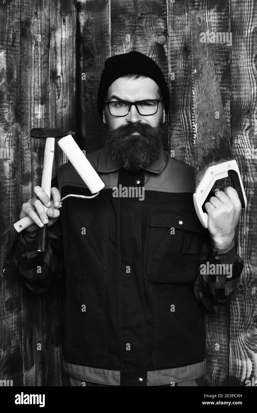 holding various building tools: saw, hammer, roller paint with serious face on brown wooden studio background. bearded painter, long beard, brutal caucasian hipster with moustache Stock Photo