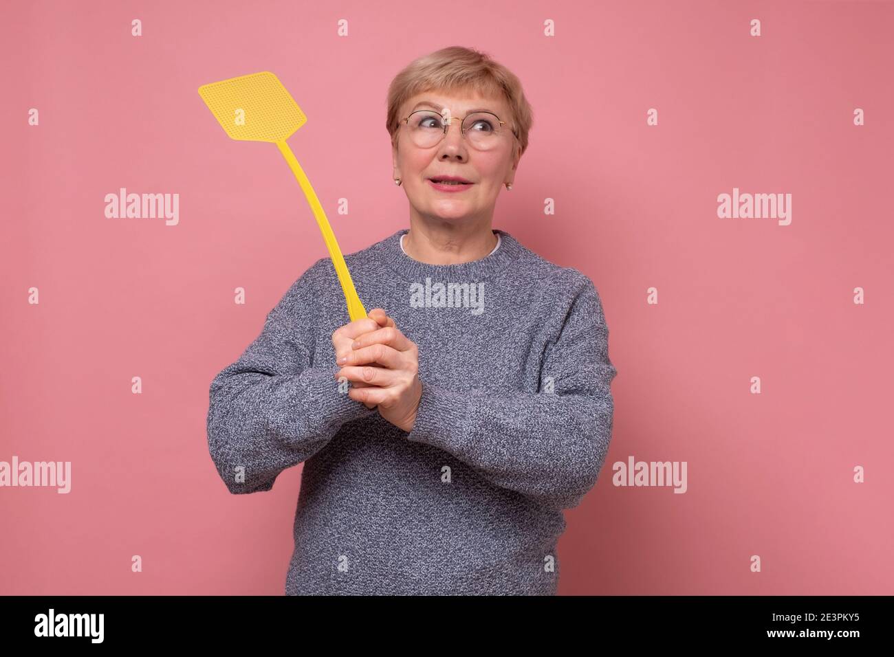 Caucasian senior woman with fly swatter trying to kill a mosquito. Studio shot on pink wall. Stock Photo