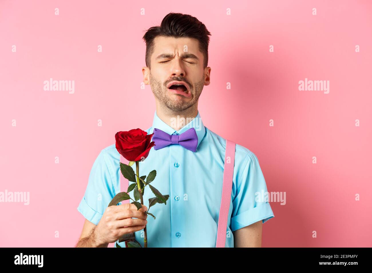 Heartbroken guy in funny bow-tie crying over girlfriend, standing alone with red rose on pink background and sobbing, break-up on Valentines day Stock Photo