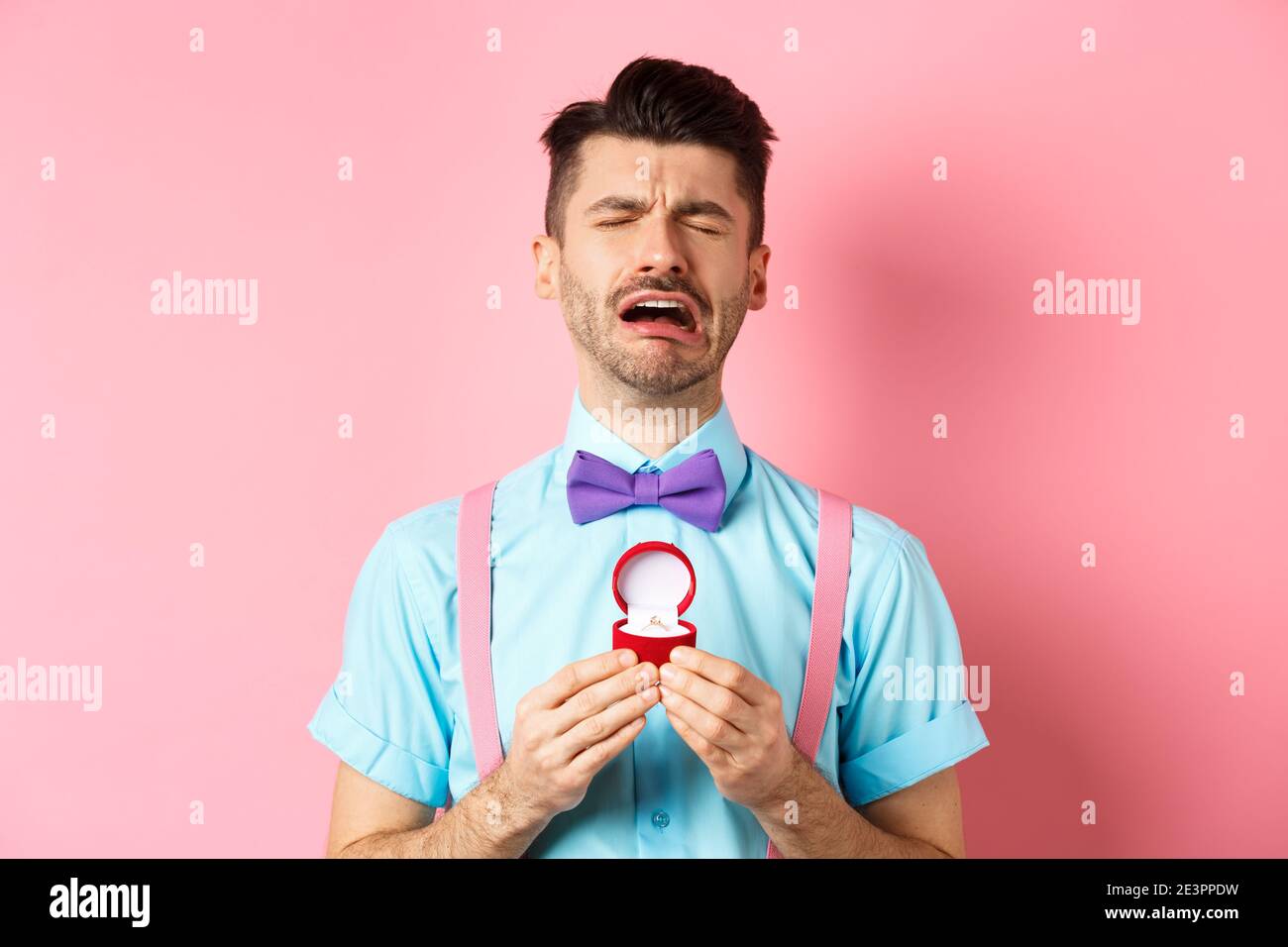 Valentines day. Heartbroken guy crying and holding engagement ring, sobbing from break-up, standing over pink background Stock Photo