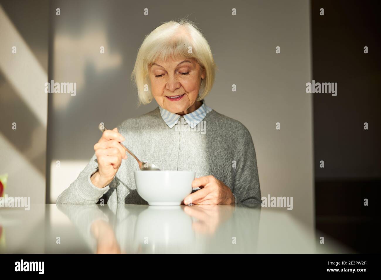 Contented elderly woman having breakfast in the sunlit kitchen Stock Photo