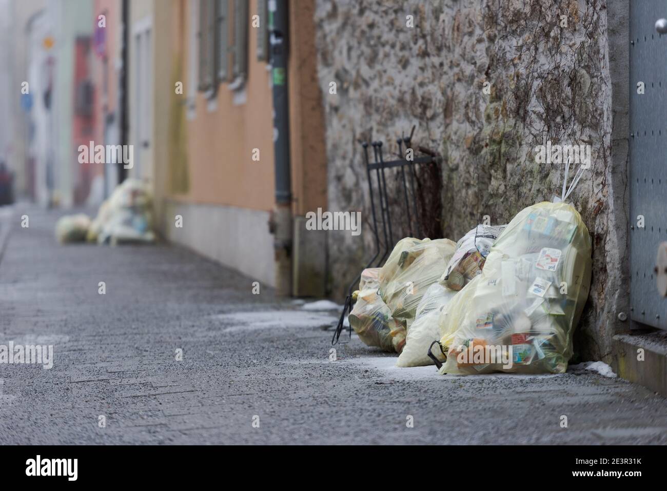 Yellow plastic sacks containing plastic garbage piled up near front-doors on sidewalk of street for waste collection in old town with stone houses Stock Photo