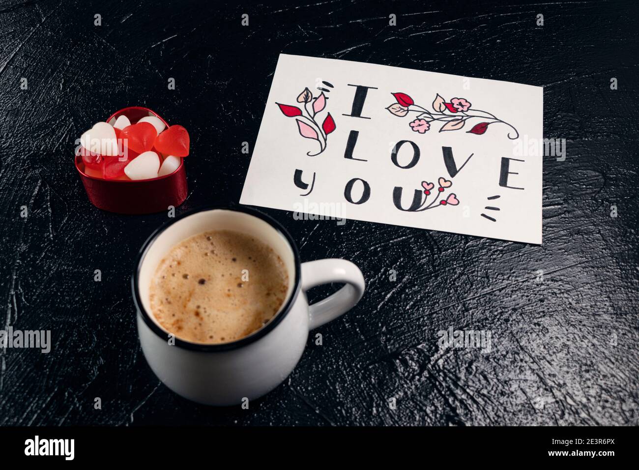 Mug of coffee, red and white heart-shaped marmalade and a valentine card with the word I love you on black background. Valentine's Day breakfast. Valentine's day concept. Soft focus Stock Photo