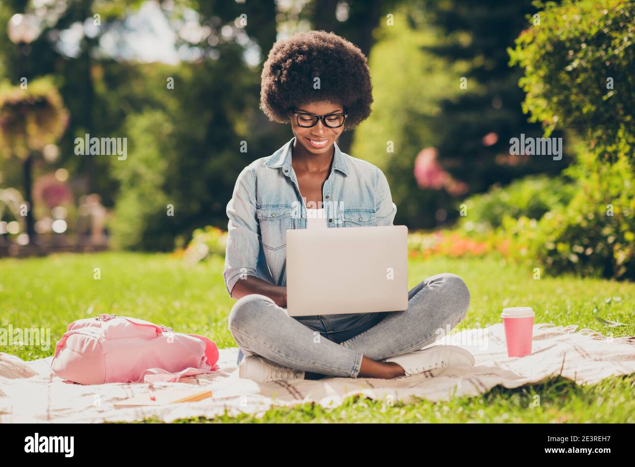 Photo portrait of black skinned young female student working with computer sitting in park wearing spectacles near bag cup of hot drink Stock Photo