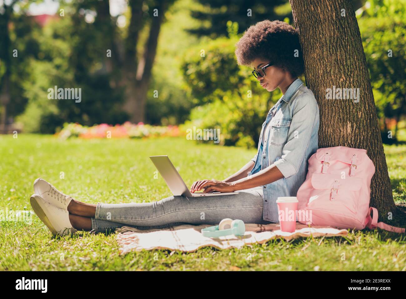 Full length body size side profile photo of black skinned woman with curly hair working on laptop browsing internet in city park Stock Photo