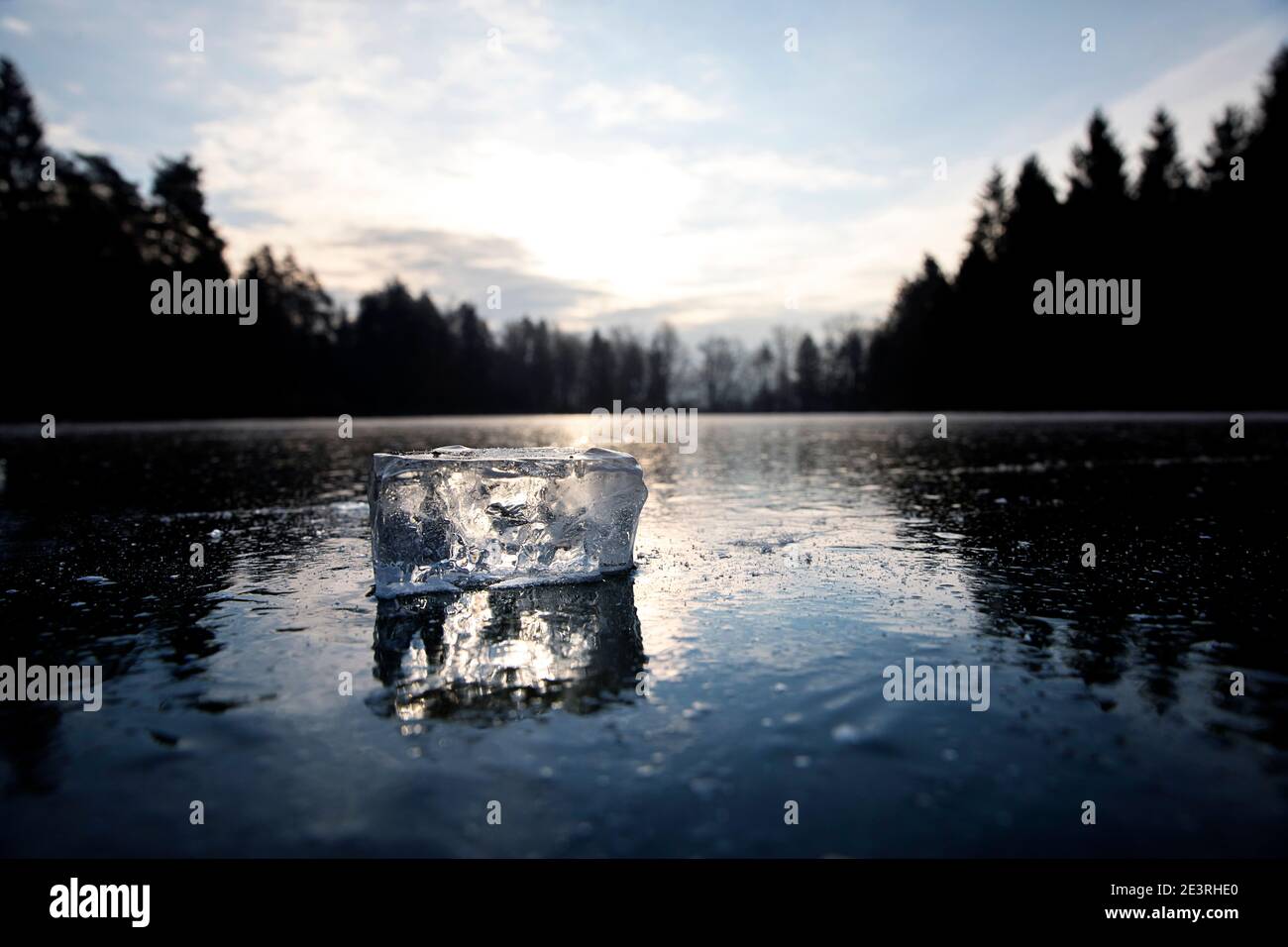 A clear cube of ice and its reflection on flat frozen lake, at sunrise, lit by sun from the back, Slovenia, Stock Photo