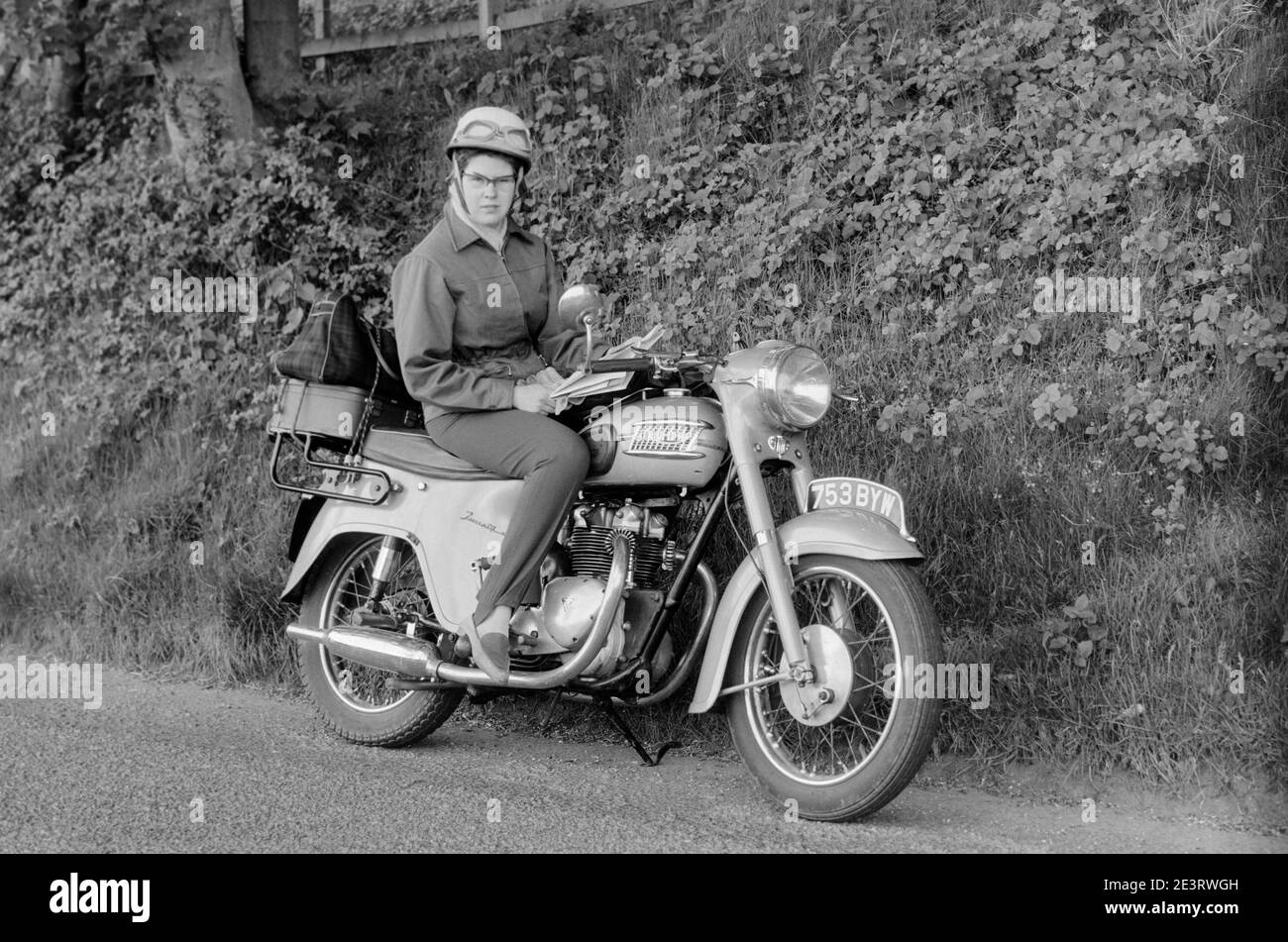Vintage late 1950s black and white photograph of a woman wearing a crash helmet, sitting on a Triumph Twenty One motorcycle, registration  number 753 BYW. The motorcycle is parked on its stand and not running. Stock Photo