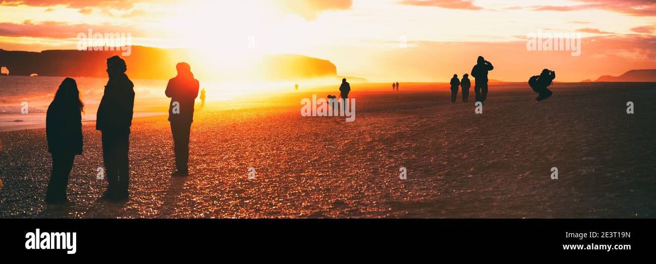 People silhouette walking on Iceland beach summer travel at sunset flare. Crowd of tourists visiting famous icelandic attraction, Reynisfjara Black Stock Photo