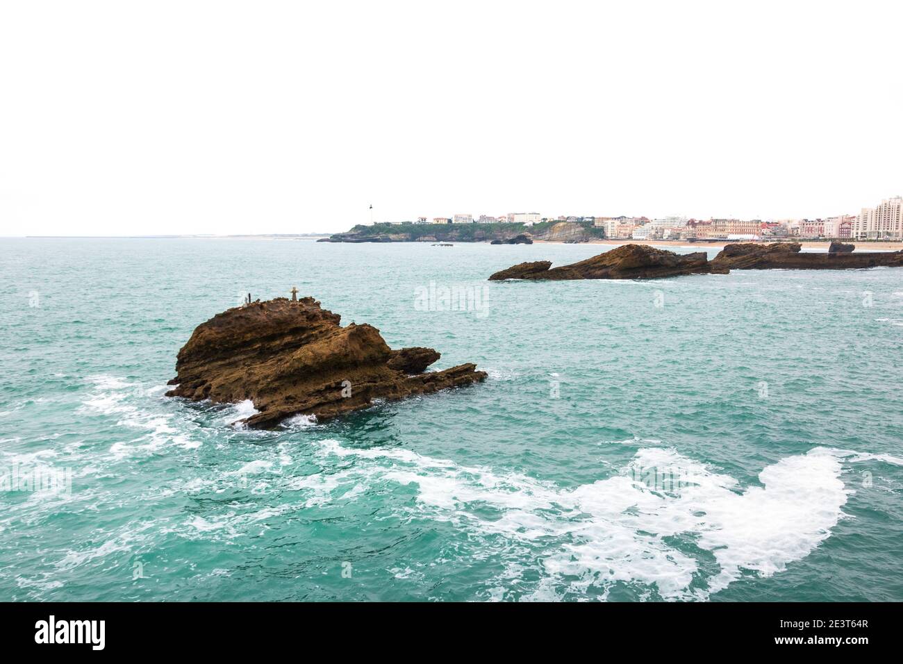 Biarritz (France). View from Rock of the Virgin Mary on rock with cross (commemorating drowned sailors) in the ocean water, city beach and lighthouse Stock Photo