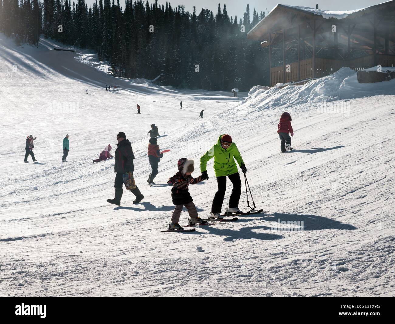 Mom teaches a child to ski among other vacationers at a ski resort on a sunny winter day. Stock Photo