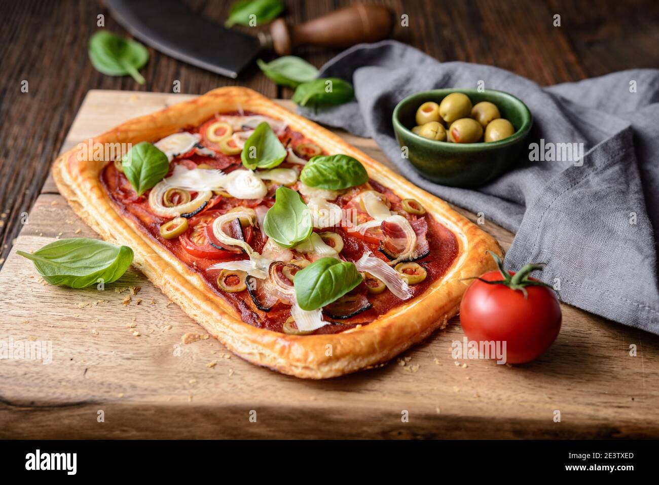 Homemade puff pastry pizza with bacon slices, tomatoes, green olives, cheese and onion, topped with basil leaves on rustic wooden background Stock Photo