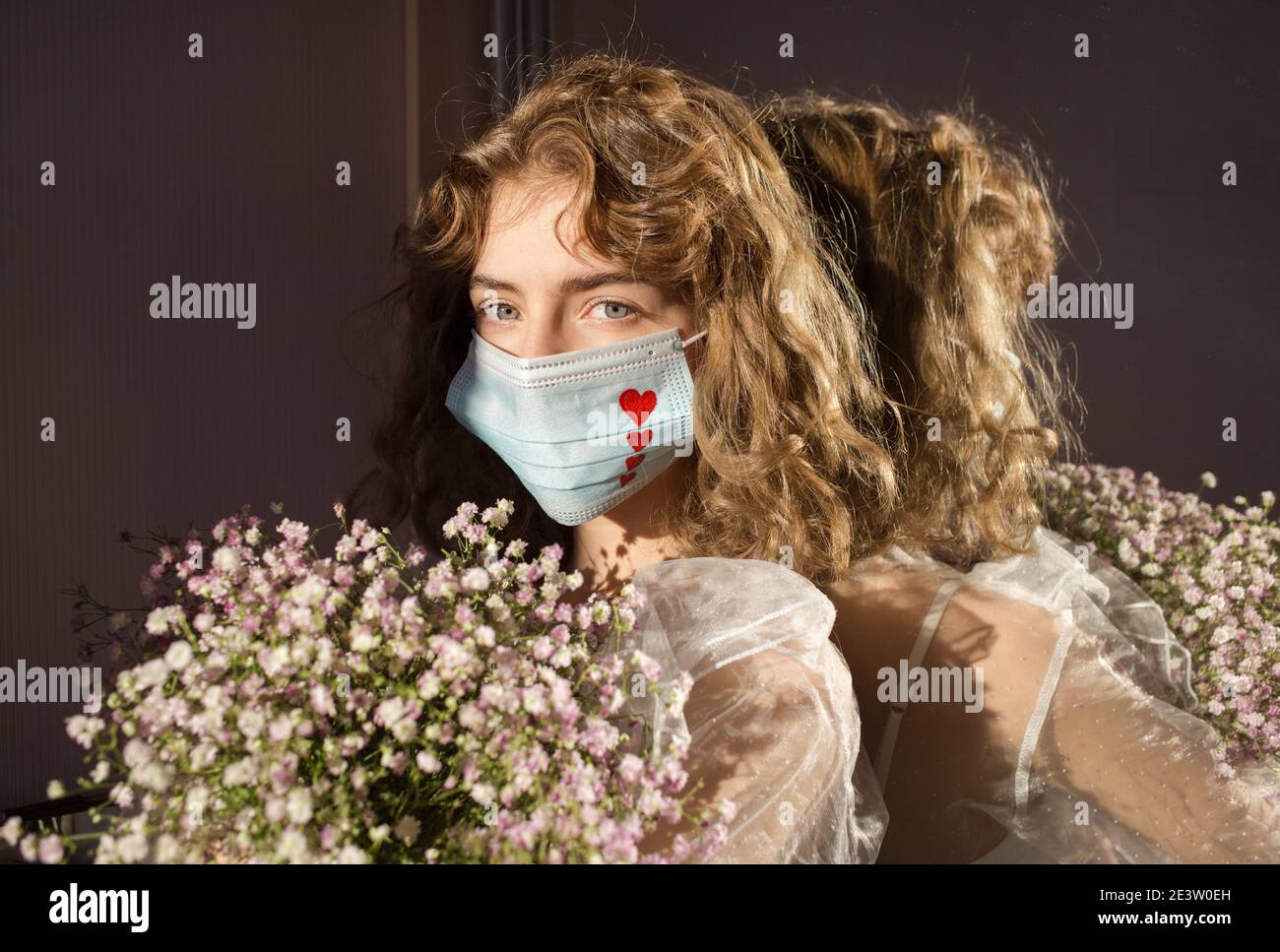 portrait of cute teenage girl in a medical mask with red hearts painted on it, with a bunch of gypsophila flowers. Reflection in the mirror. valentine Stock Photo