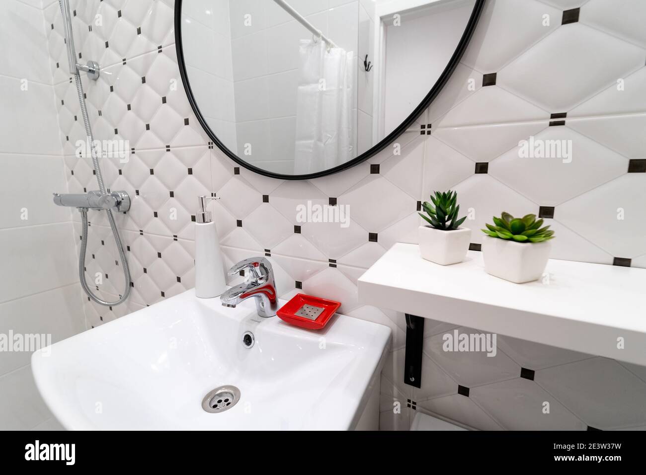 Modern white and black toilet interior. White sink with water tap and toiletries, above it large round mirror on the wall of the ceramic tiles Stock Photo