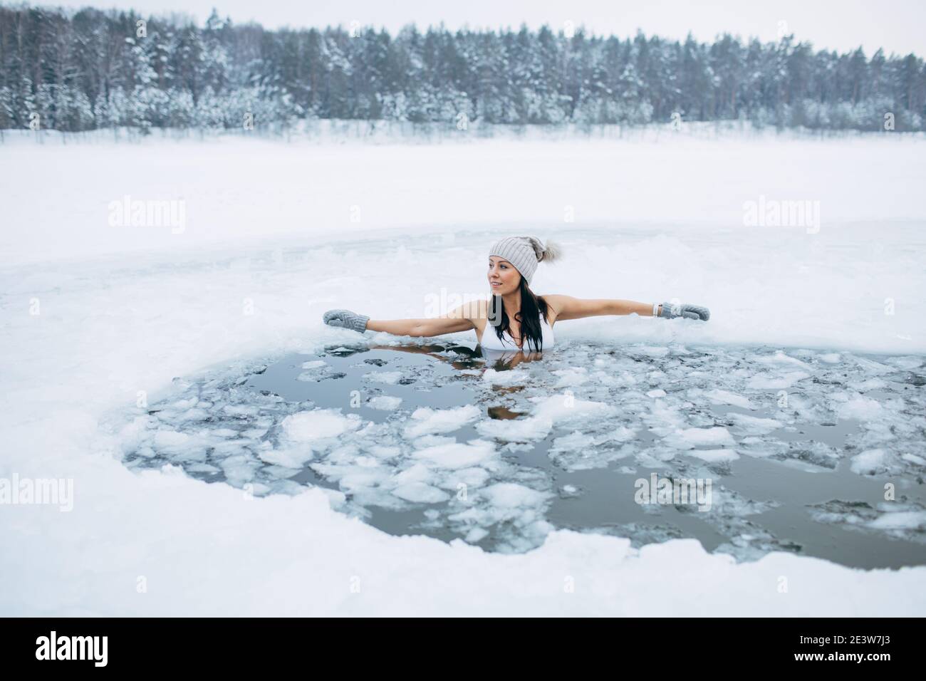 Winter swimming. Woman in frozen lake ice hole. How to swim in cold water. Beautiful young female smiling. Hat and gloves swimming clothes. Stock Photo