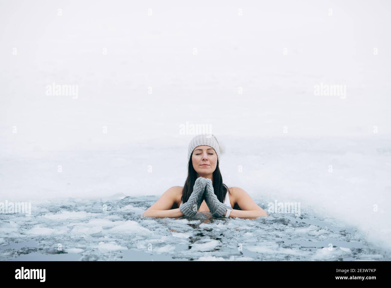 Winter swimming. Woman in frozen lake ice hole. How to swim in cold water. Beautiful young female in zen meditation. Hat and gloves swimming clothes. Stock Photo