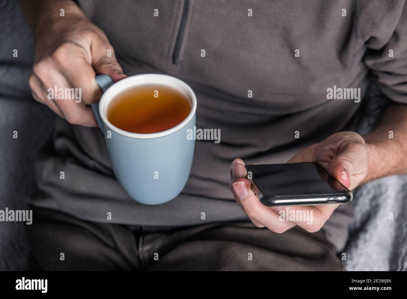 Male hands are holding a mobile phone and a cup of tea. Close-up. Man sitting at home on the couch drinking tea and texting with friends  on the phone Stock Photo