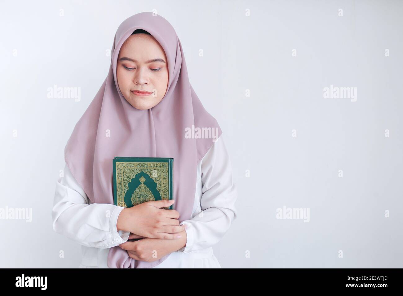 Young Asian Islam woman wearing headscarf is holding holy al quran with close eyes and calm face. Indonesian woman on gray background. Stock Photo