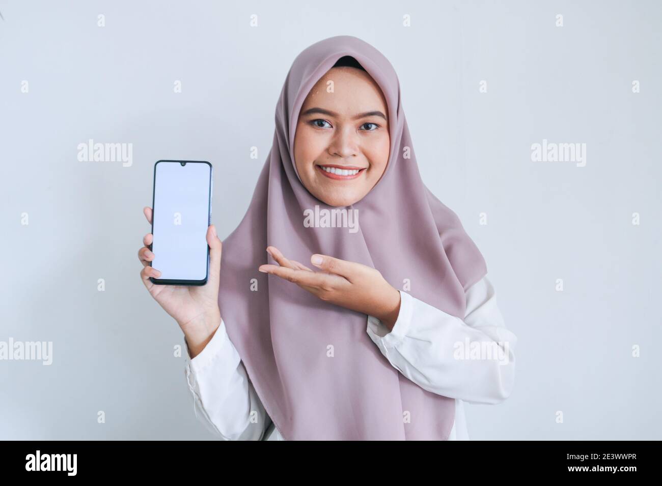 Young Asian Islam woman wearing headscarf is pointing finger on white screen of phone with smile and happy feeling. Indonesian woman on gray backgroun Stock Photo