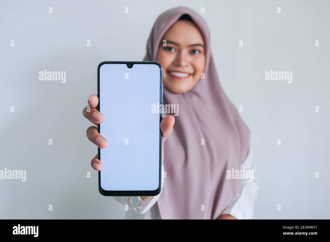 Young Asian Islam woman wearing headscarf is pointing finger on white screen of phone with smile and happy feeling. Indonesian woman on gray backgroun Stock Photo