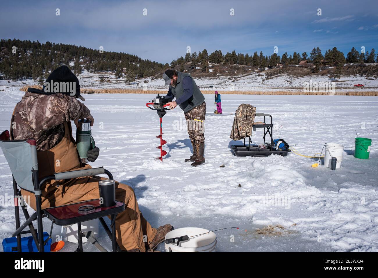 Three people ice fishing on a frozen lake. One sits in a chair pouring  coffee, a man drills a hole in the ice, and a woman fishes with a pink pole  Stock