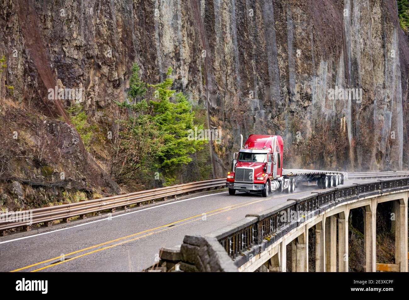Big rig red semi truck with chrome pipes transporting empty flat bed semi trailer running on the winding wet road with rain dust driving on the bridge Stock Photo
