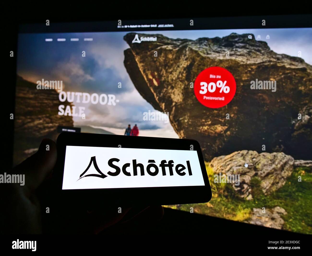 Person holding smartphone with logo of outdoor clothing company Schöffel on screen in front of online shop website. Focus on mobile phone monitor. Stock Photo