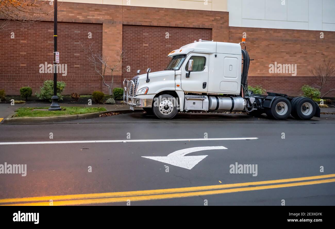 Industrial grade low cab white big rig semi truck tractor with pipes grille guard and without semi trailer standing on wet after rain city street park Stock Photo