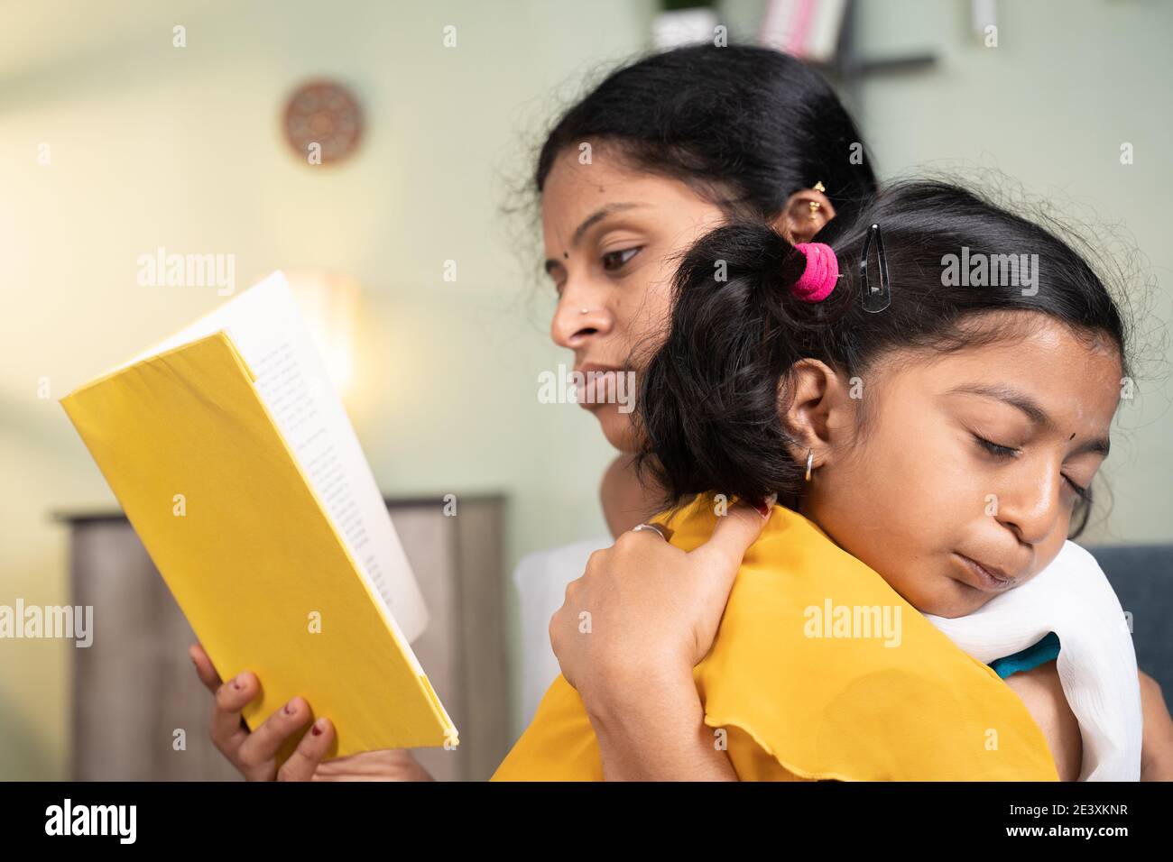 Girl kid sleeping on Mother's shoulder while mother busy reading book and making her kid sleep at home Stock Photo