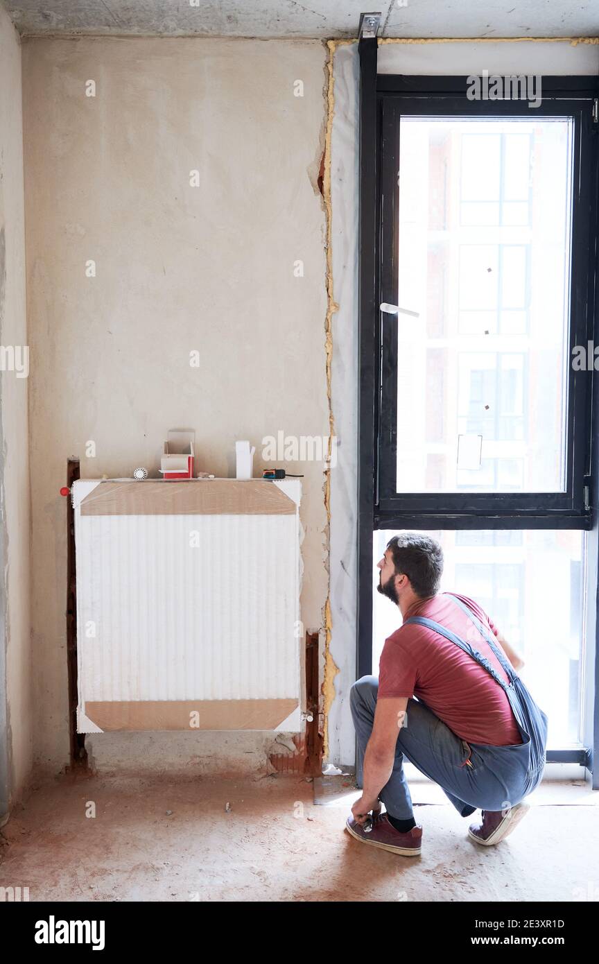 Plumber installing water radiator in new empty room next to window. Fitting lower valve. Male worker during installation process. Construction, maintenance, home refurbishment concept Stock Photo