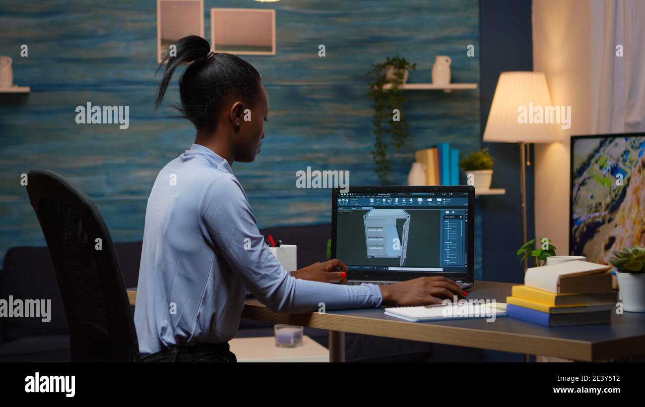 African lady enginer analysing digital mechanism of container checking prototype of project looking on laptop sitting at desk in living room office late night. Black employee using modern technology Stock Photo