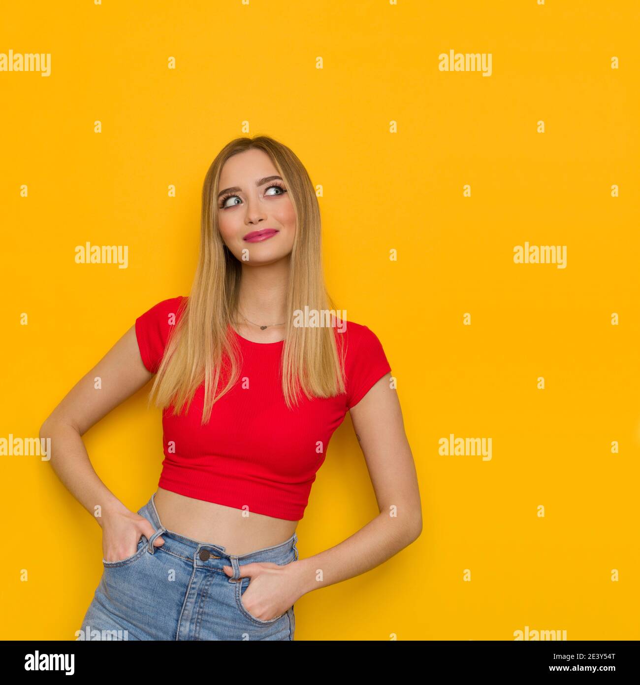 Smiling young woman in red top is holding hands in pockets and looking at the side. Front view. Waist up studio shot on yellow background. Stock Photo