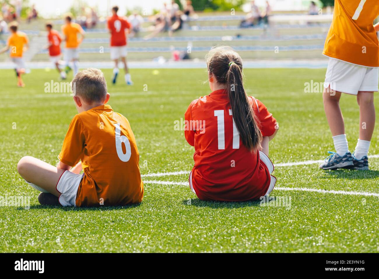Young boy and girl in a sports team. Happy kids playing sports on summer sunny day. Teammates sitting on grass soccer venue. Young players watching to Stock Photo