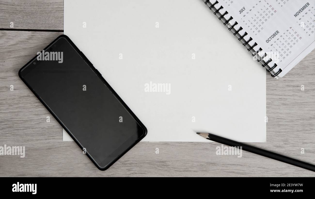 Top view of workplace. Empty paper blank, spiral calendar, black pencil and phone on wooden table. Stock Photo