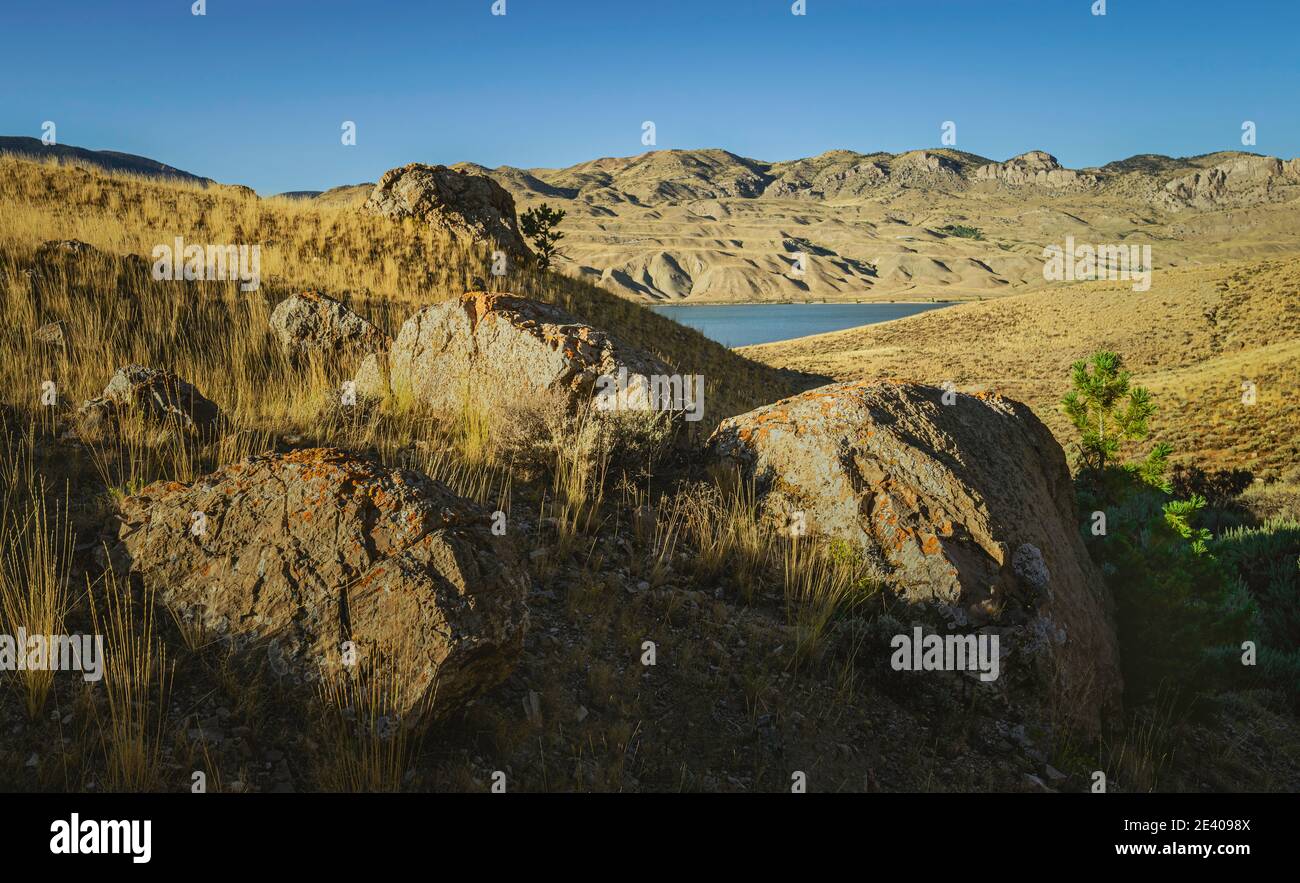 Arid landscape  with boulders, grasses, and foothills of the Rockies with glimpse of reservoir under blue sky near Cody, Wyoming, USA. Stock Photo