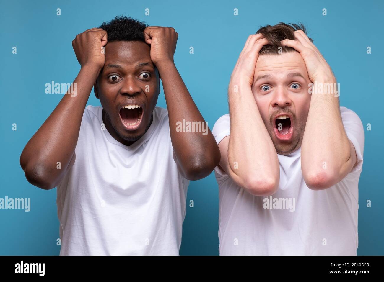 African and caucasian men feel scared afraid, confused and shocked by unexpected news. Studio shot on blue wall Stock Photo