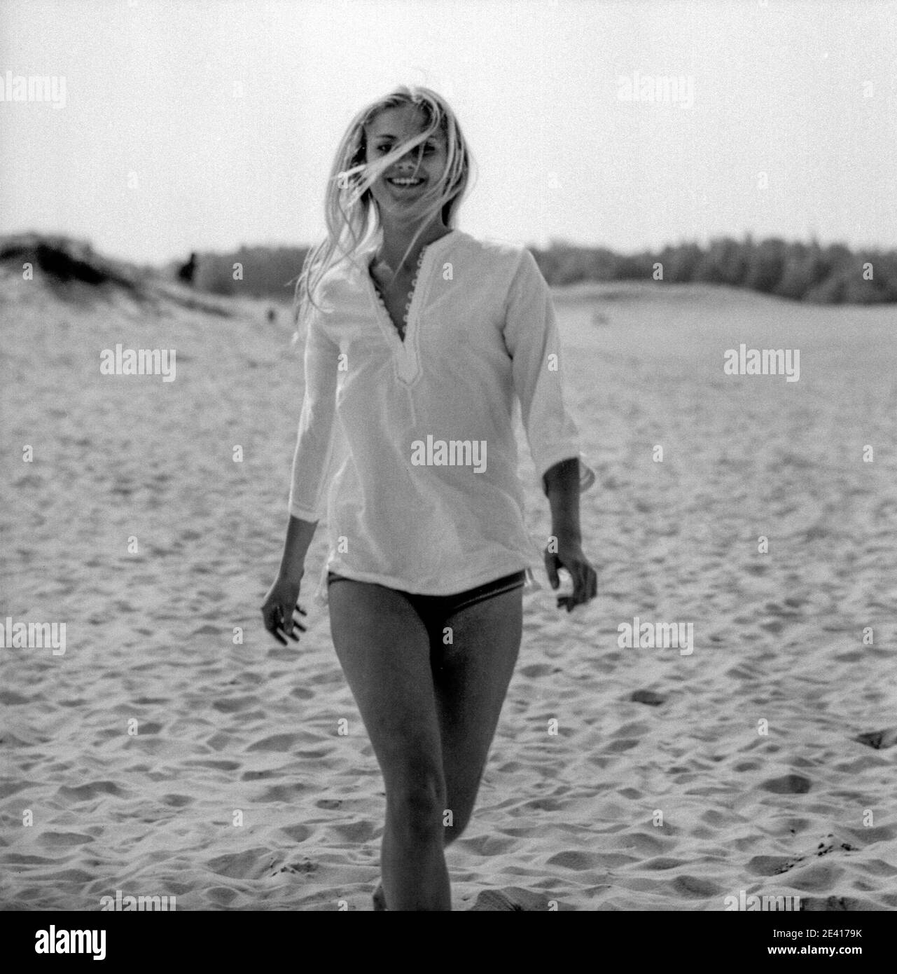 A young smiling blonde woman strolls towards the camera on a deserted beach in the 1960s Stock Photo