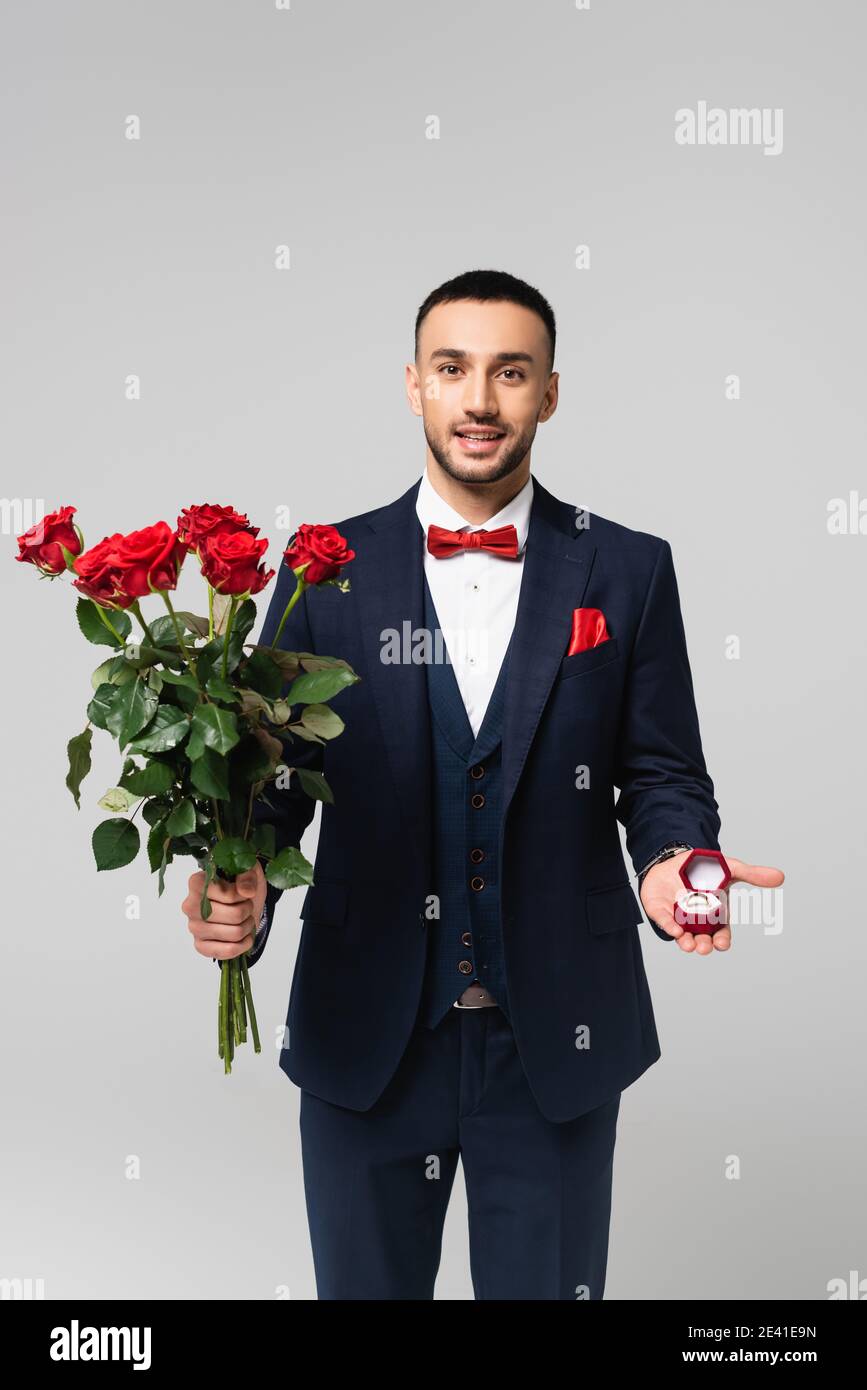 happy hispanic man in elegant suit holding red roses and jewelry box with wedding ring isolated on grey Stock Photo